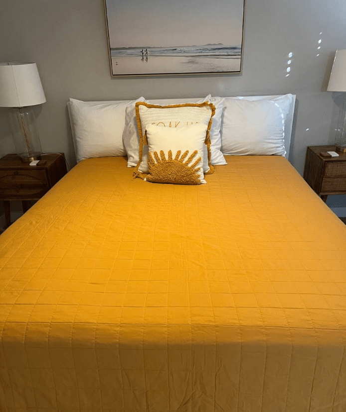 Bright yellow quilt covers a neatly made bed with white pillows and a sun-themed decorative pillow. Room includes bedside lamps and art.